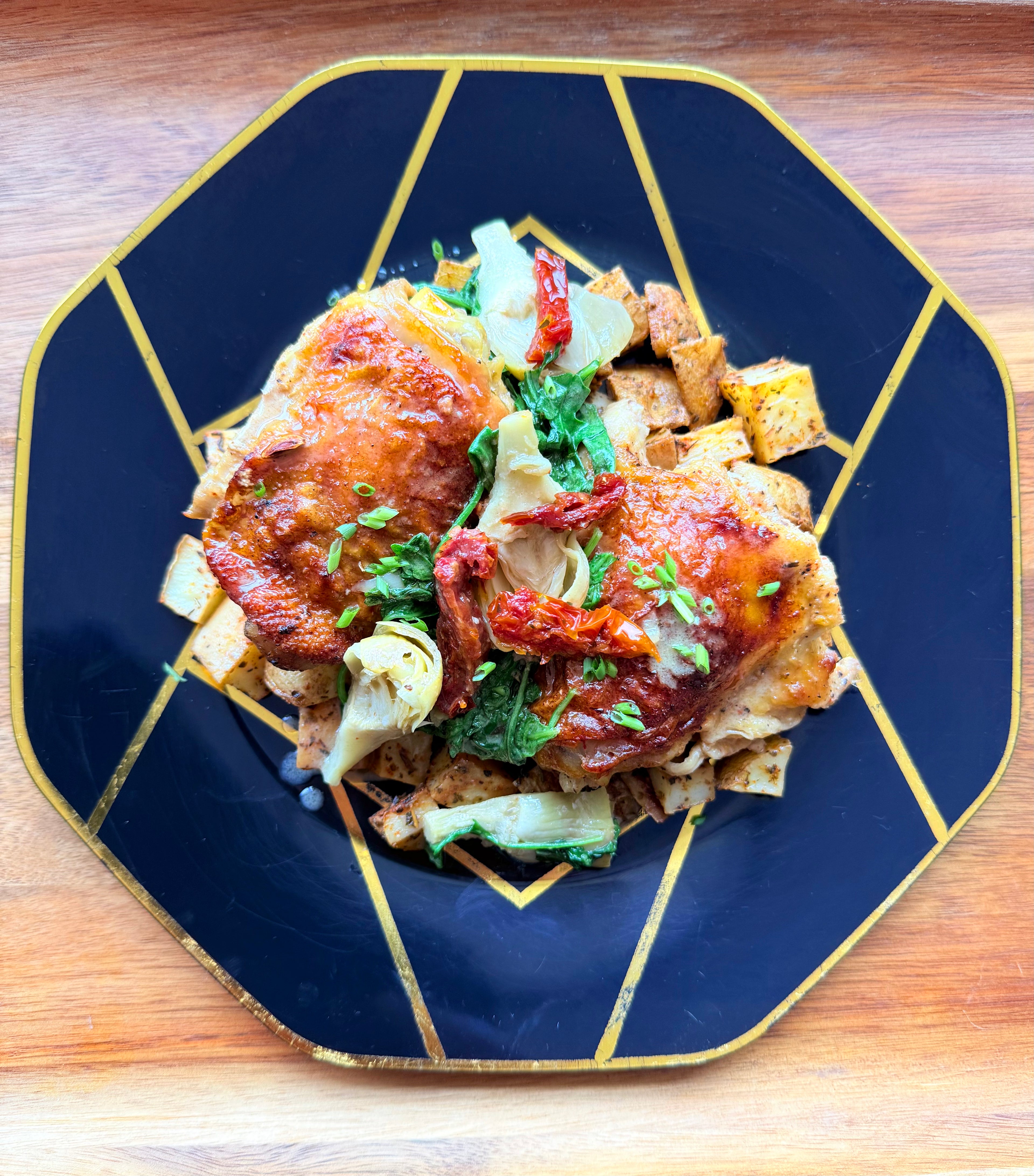 Two glazed Tuscan braised chicken thighs with sun-dried tomatoes, artichokes, and herbs on a navy and gold octagonal plate.
