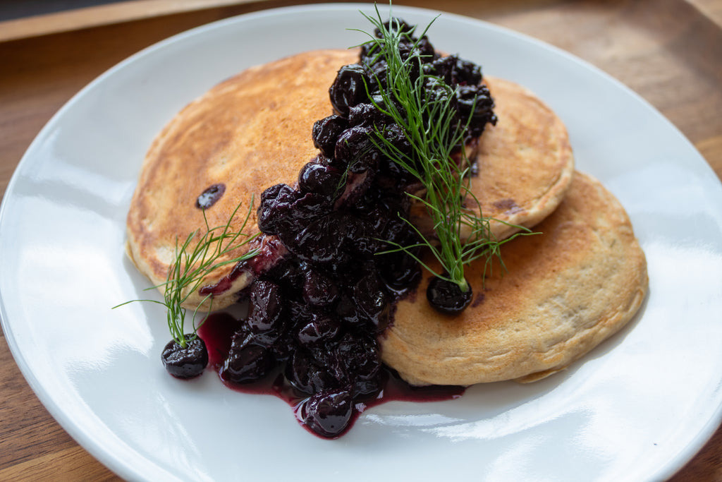 Two golden Kodiak pancakes topped with rich blueberry compote and fresh dill, served on a white plate.