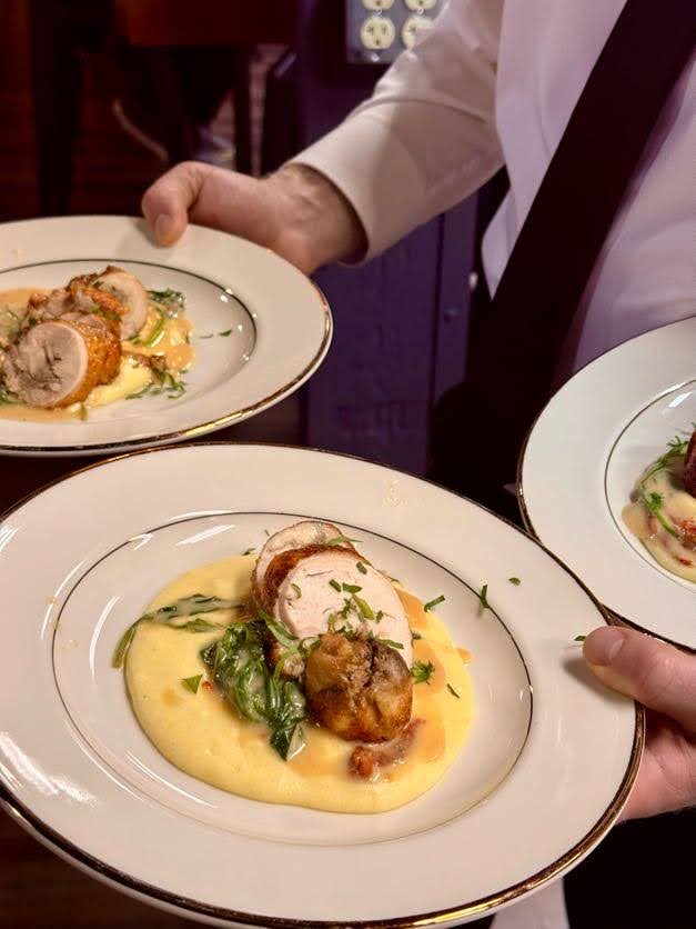 Roasted garlic braised chicken thighs plated on creamy polenta with asparagus, garnished with chives, served on gold-rimmed
