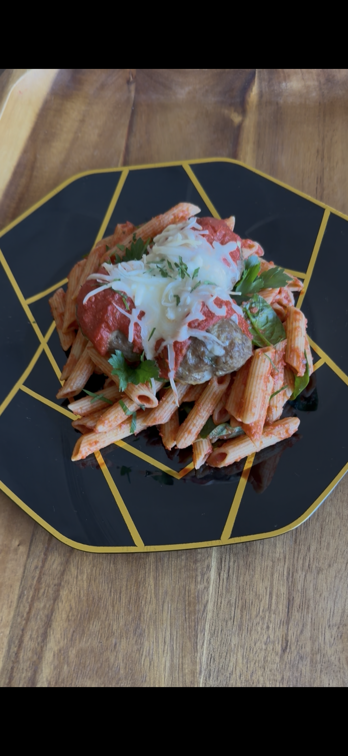 Penne pasta topped with a wagyu meatball, marinara sauce, melted shredded cheese, and fresh parsley on a black geometric